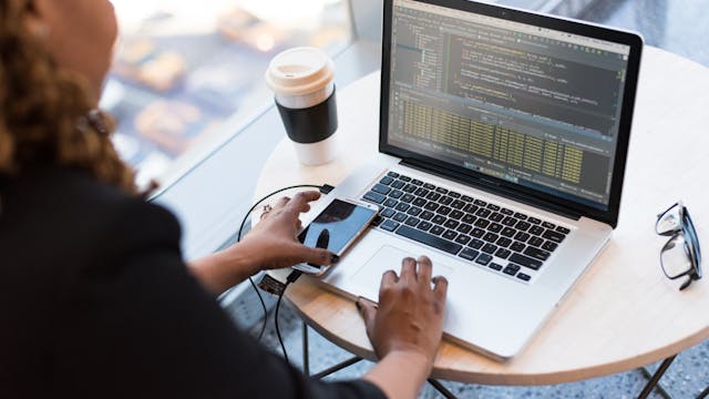 Black woman programming on a laptop with coffee, smartphone, and glasses on a desk in an office.